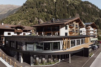 Sommeransicht des Familienhotels Kindl in den Tiroler Alpen mit umliegenden Wiesen, Bergen und traditioneller Architektur.