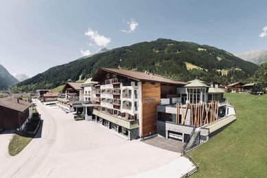 Sommeransicht des Familienhotels Kindl in den Tiroler Alpen mit umliegenden Wiesen, Bergen und traditioneller Architektur.