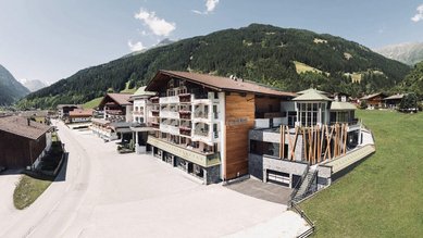 Sommeransicht des Familienhotels Kindl in den Tiroler Alpen mit umliegenden Wiesen, Bergen und traditioneller Architektur.