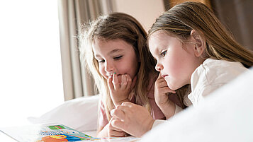 Zwei Mädchen liegen auf dem Bett in ihrem Familienzimmer im Familienhotel und schauen ein Buch an.
