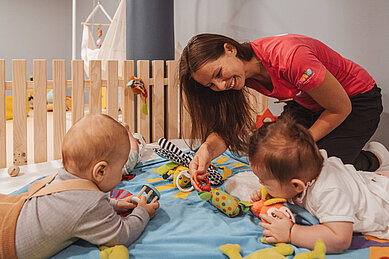 Zwei Babys liegen gemeinsam mit einer Babybetreuerin des Familienhotels Huber in Südtirol auf einer Spieldecke und beschäftigen sich mit altersgerechten Spielsachen.