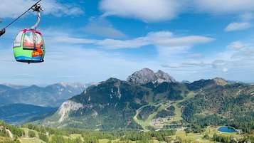 Bunte Gondel mit Familienhotel Ramsi Logo schwebt vor beeindruckender Alpenkulisse über grüne Berghänge in Kärnten