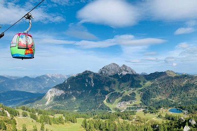Bunte Gondel mit Familienhotel Ramsi Logo schwebt vor beeindruckender Alpenkulisse über grüne Berghänge in Kärnten