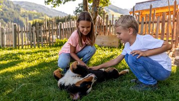 Salzburger Land mit Tieren erleben. Zwei Kinder streicheln einen jungen Hund, der auf dem Rücken liegt. Im Hintergrund ein Bauernhof in Mühlbach.
