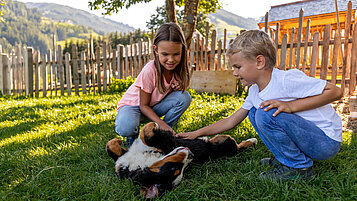 Salzburger Land mit Tieren erleben. Zwei Kinder streicheln einen jungen Hund, der auf dem Rücken liegt. Im Hintergrund ein Bauernhof in Mühlbach.