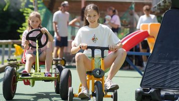 Kinder fahren lachend mit Dreirad und Gokart auf dem Spielplatz des Familienhotel Sonne Bezau.