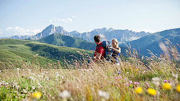 Auf einer Blumenwiese sitzen zwei Menschen und bewundern den Ausblick. Die Berge in Südtirol sind ein Grund dort wandern zu gehen.