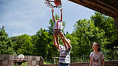 Vater hält seine Tochter in die Luft, damit diese einen Basketball auf dem Basketballplatz des Kinderhotels Bruckwirt in den Korb werfen kann.