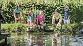 Im Familienhotel Alpenhof Dolomit Family in Südtirol können Kinder mit dem Naturdetektiv vieles entdecken. Die Kinder sitzen am Teich und beobachten eine Ente.