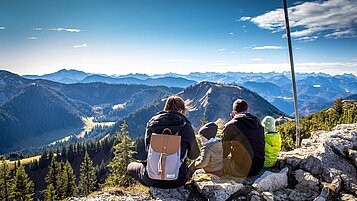 Eltern sitzen mit ihren beiden Kindern am Gipfel eines bayerischen Berges und genießen die Aussicht.