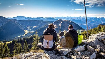 Eltern sitzen mit ihren beiden Kindern am Gipfel eines bayerischen Berges und genießen die Aussicht.