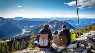 Eltern sitzen mit ihren beiden Kindern am Gipfel eines bayerischen Berges und genießen die Aussicht.