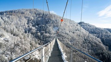 Lange Hängebrücke über einem verschneiten Waldtal an einem klaren, sonnigen Wintertag.