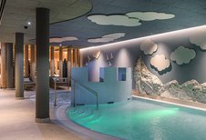 Kinderfreundlicher Indoor-Pool im Zauchenseehof mit beleuchtetem Wasser, spielerischer Burgwand, Felskulisse und Wandmotiven aus Wolken und Vögeln.