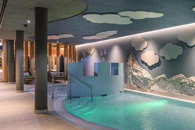 Kinderfreundlicher Indoor-Pool im Zauchenseehof mit beleuchtetem Wasser, spielerischer Burgwand, Felskulisse und Wandmotiven aus Wolken und Vögeln.