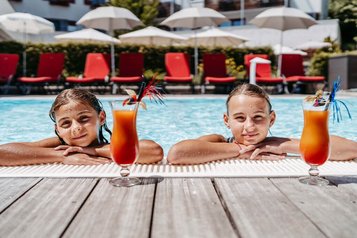 Zwei Mädchen genießen bunte Kinder-Cocktails am Poolrand des Familienhotels Amiamo an einem sonnigen Sommertag.