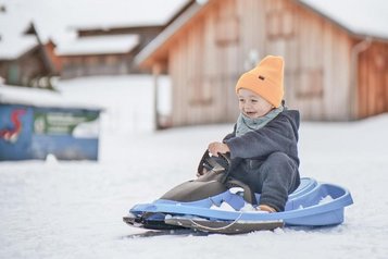 Ein Kleinkind sitzt auf einem Schlitten im Schnee beim Winterurlaub am Familienhotel Gorfion in Liechtenstein.