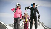 Familie mit Kind geht in Skiausrüstung und mit Skiern auf den Schultern in Richtung Skigebiet.