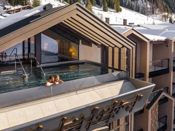 Außenansicht des Infinity-Pools des Hotels Zauchenseehof, in dem zwei Personen im warmen Wasser entspannen; der Pool ist in eine moderne Dachkonstruktion integriert und bietet einen Panoramablick auf die verschneiten Berge und Skipisten.