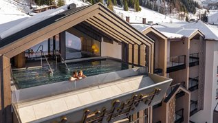 Außenansicht des Infinity-Pools des Hotels Zauchenseehof, in dem zwei Personen im warmen Wasser entspannen; der Pool ist in eine moderne Dachkonstruktion integriert und bietet einen Panoramablick auf die verschneiten Berge und Skipisten.