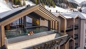 Außenansicht des Infinity-Pools des Hotels Zauchenseehof, in dem zwei Personen im warmen Wasser entspannen; der Pool ist in eine moderne Dachkonstruktion integriert und bietet einen Panoramablick auf die verschneiten Berge und Skipisten.