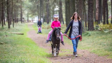 Eine Frau hält ein Pony an den Zügeln, auf dem ein Mädchen reitet.
