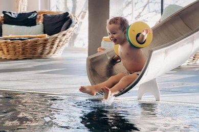 Kleinkind rutscht fröhlich die Wasserrutsche im Indoorpool im Familienhotel Amiamo im Salzburger Land hinunter.