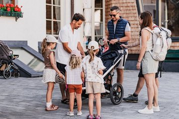  Zwei Familien mit Kindern und Kinderwagen bereiten sich vor dem Familienhotel Tirolerhof auf eine gemeinsame Wanderung vor.