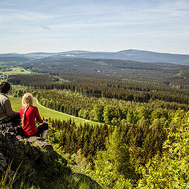 Blick vom Kammerweg vom Bärensteig ins grüne Tal. Ein Paar sitzt auf einen Felsen und genießt die Aussicht.