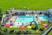 Bade- und Wellnessoase im Familienhotel Egger mit mehreren Außenpools, Wasserrutschen, Liegen und Sonnenschirmen inmitten grüner Natur.