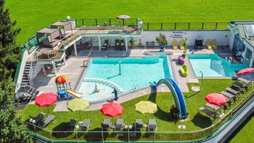 Bade- und Wellnessoase im Familienhotel Egger mit mehreren Außenpools, Wasserrutschen, Liegen und Sonnenschirmen inmitten grüner Natur.