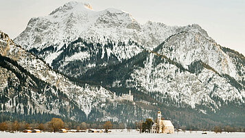 Bayern im Winter: schneebedecktes Bergpanorama mit leichtem Nebel und einer Kapelle im Vordergrund