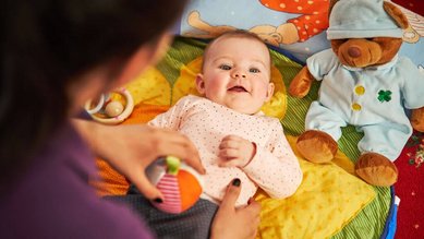 Ein Baby liegt fröhlich auf einer bunten Spieldecke, umsorgt von einer Betreuerin in der liebevollen Babybetreuung im Familienhotel Amiamo.