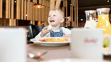 Ein Baby sitz am Tisch beim Essen und lacht und hat ein Stück Brot in der Hand.