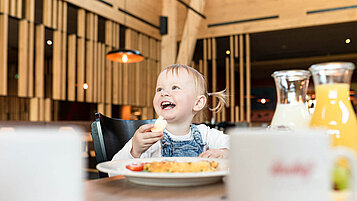 Ein Baby sitz am Tisch beim Essen und lacht und hat ein Stück Brot in der Hand.