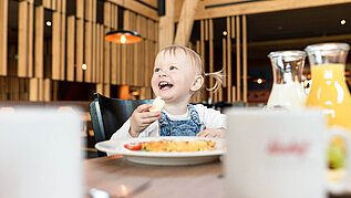 Ein Baby sitz am Tisch beim Essen und lacht und hat ein Stück Brot in der Hand.