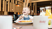 Ein Baby sitz am Tisch beim Essen und lacht und hat ein Stück Brot in der Hand.