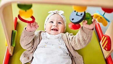 Glückliches Baby spielt mit einem Spielebogen im Babyspielbereich des Familienhotels Aigo welcome family in Oberösterreich.