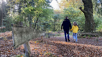 Ein Papa mit Tochter wandert über den mit Laub bedeckten Waldboden.