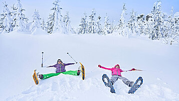 Zwei Frauen liegen im Tiefschnee mit Scheeschuhen an den Füßen.