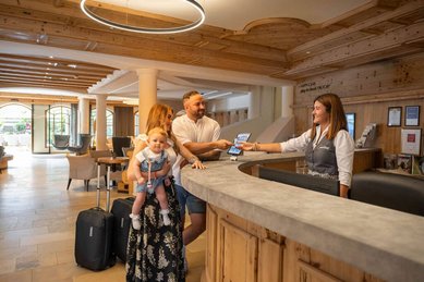Familie mit Kleinkind beim Check-in an der Rezeption des Familienhotels Alpenhotel Kindl mit freundlicher Mitarbeiterin.