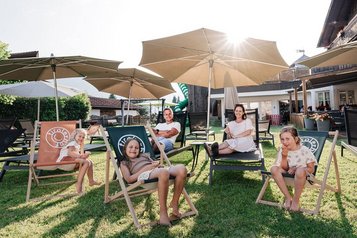 Eine Familie entspannt gemeinsam im Garten des Familienhotels Tirolerhof unter großen Sonnenschirmen auf Liegestühlen.