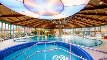 Helles Hallenbad mit großem Rundbecken und Whirlpool im Vordergrund, Holzdecke mit runder Lichtkuppel und Liegen entlang der Fensterfront.