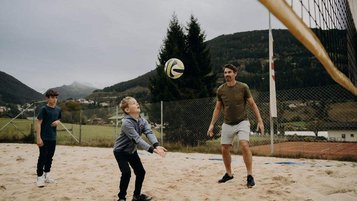 Eine Familie spielt auf einem Sandplatz vor bergiger Kulisse Beachvolleyball im Außenbereich des Hotel Sonngastein im Salzburger Land.