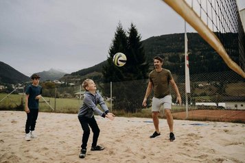 Eine Familie spielt auf einem Sandplatz vor bergiger Kulisse Beachvolleyball im Außenbereich des Hotel Sonngastein im Salzburger Land.