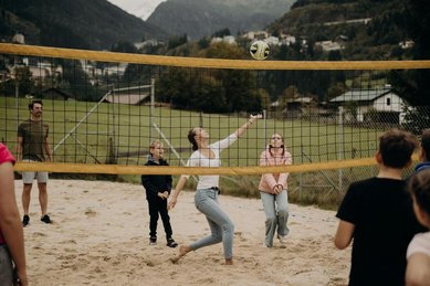 Eine Familie spielt auf einem Sandplatz vor bergiger Kulisse Beachvolleyball im Außenbereich des Hotel Sonngastein im Salzburger Land.