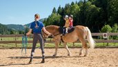 zwei Mädchen sitzen auf einem Pferd am Reitplatz des Spa- & Familien-Resorts Krone unter Beobachtung einer Trainerin.. 