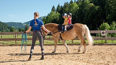 zwei Mädchen sitzen auf einem Pferd am Reitplatz des Spa- & Familien-Resorts Krone unter Beobachtung einer Trainerin.. 