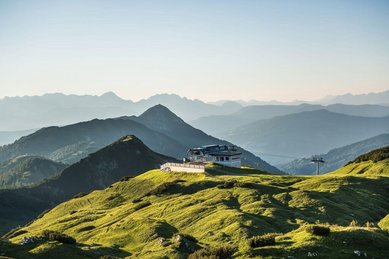 Panoramablick auf eine Berglandschaft mit einer Berghütte und Seilbahnstation auf einem grünen Gipfel
