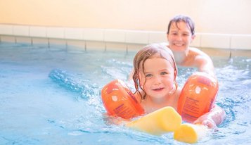 Ein fröhliches Kleinkind lacht, während es im Schwimmbad mit seiner Mutter spielt, beide umgeben von klarem Wasser und einer farbenfrohen Poolumrandung, was die familiäre und kinderfreundliche Atmosphäre im Familienhotel Deichkrone unterstreicht.